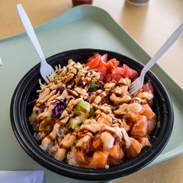 A poke bowl with salmon, avocado, cabbage, and sauces, served with plastic forks on a tray.
