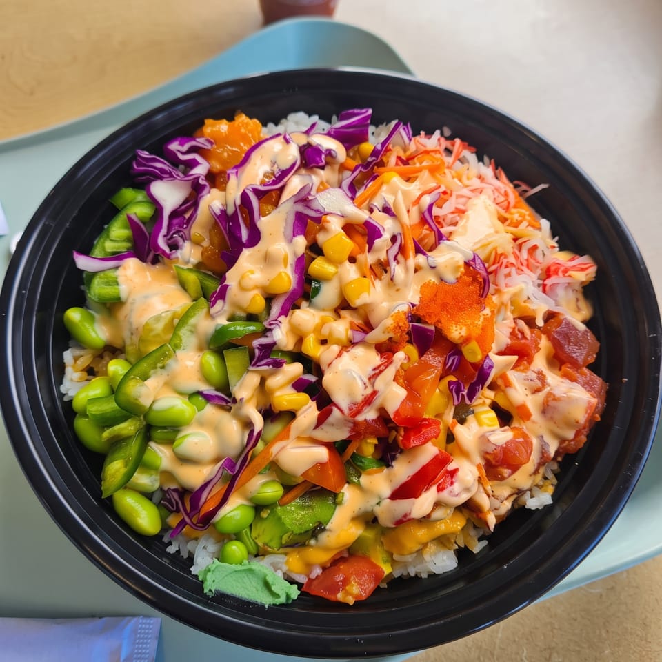 Colorful poke bowl with rice, edamame, corn, red cabbage, and various toppings drizzled with sauce.